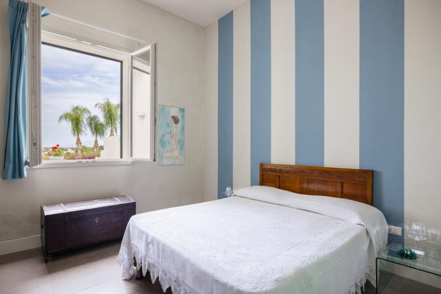 Bright and cozy bedroom with striped blue and white walls, an antique wooden chest, and an open window with a view in a Sicily luxury villa