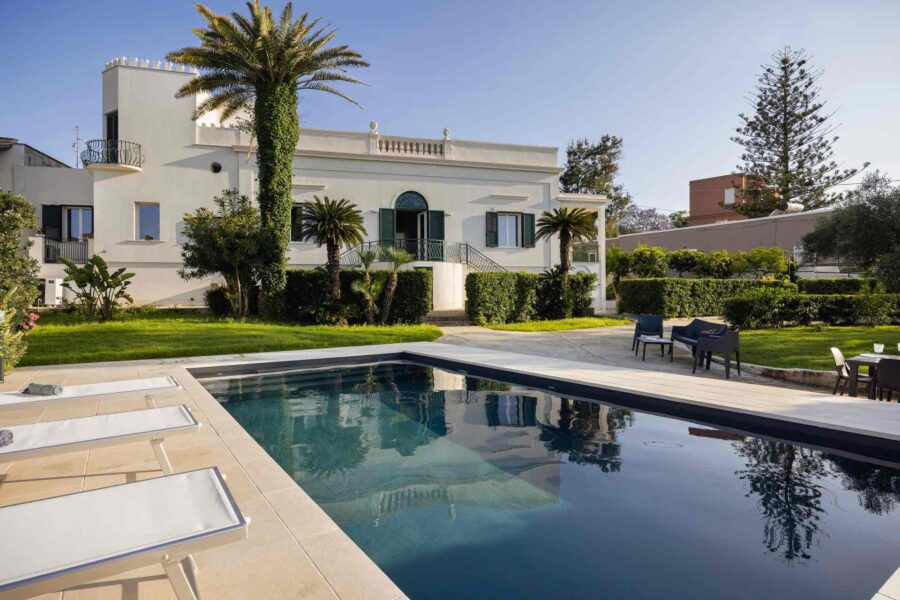 Luxury Sicilian villa with pool surrounded by lush greenery and palm trees.
