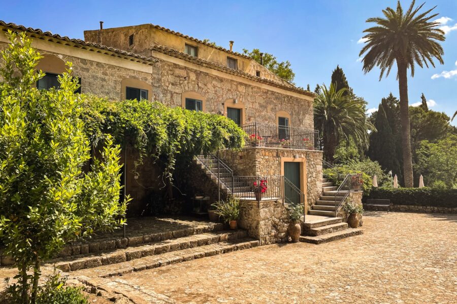 Stone cottage in Sicily with a spacious courtyard, lush greenery, and a backdrop of palm trees under a bright blue sky.