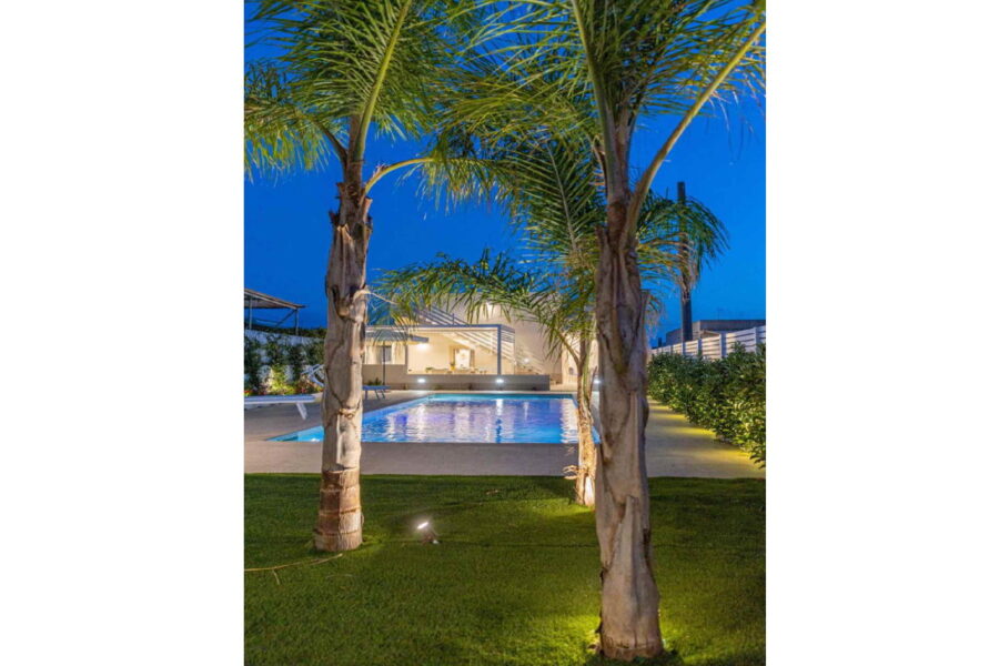 Villa Blue Oasis in Sicily at night, featuring a beautifully lit private pool surrounded by lush greenery and palm trees.