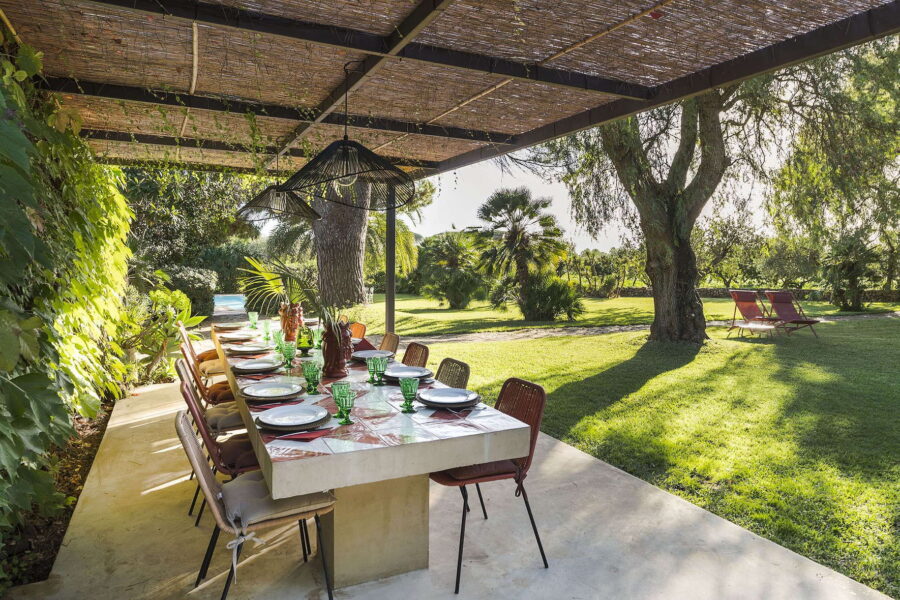 Long outdoor dining table with ceramic tiles, shaded pergola, and garden views at Villa Selvira, one of the most picturesque Sicily villas with pool.