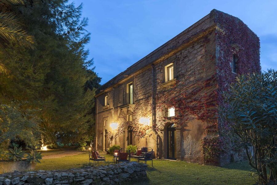 Stone facade of Villa Selvira illuminated at dusk, with ivy-covered walls, warm garden lighting, and outdoor seating – a standout Sicily villa with pool.