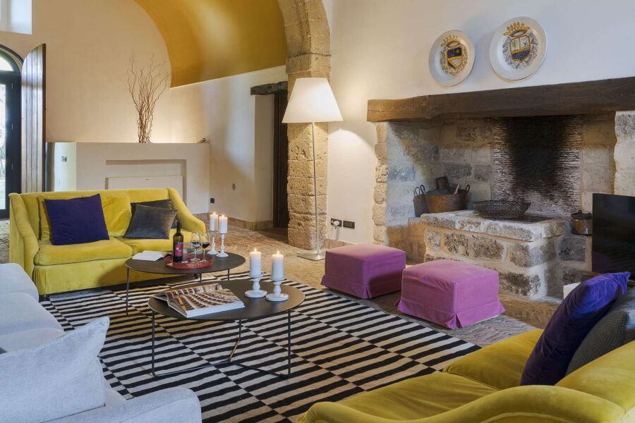 Stylish living room of Villa Selvira, a Sicily luxury villa, featuring vibrant yellow sofas, striped rugs, and a stone fireplace.